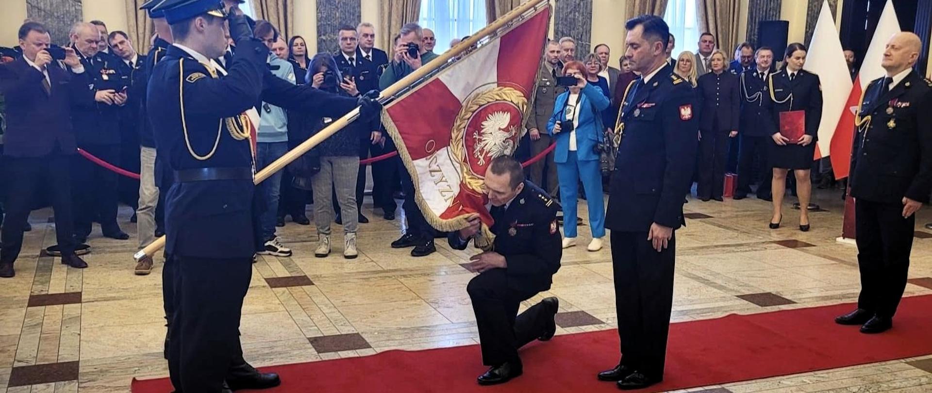 W sali pałacowej, na czerwonym dywanie stoją strażacy. Strażak z pocztu sztandarowego pochyla sztandar, jeden ze strażaków klęczy i całuje płat, drugi stoi obok. Wokół przyglądają się temu goście.