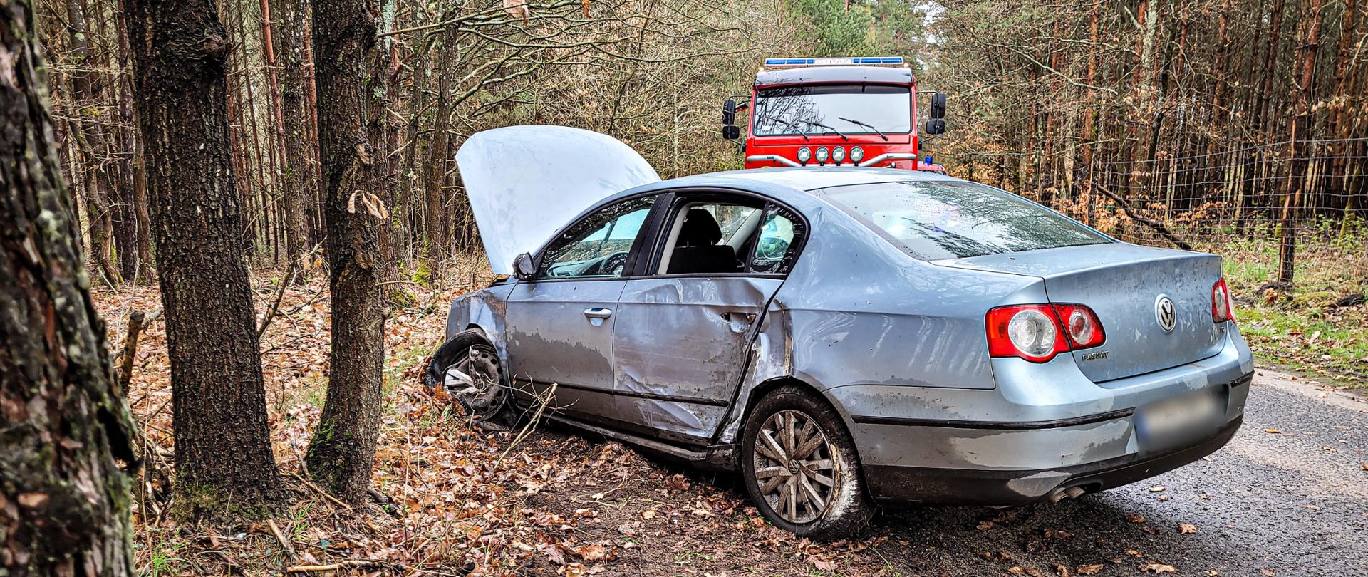 W środkowej części zdjęcia znajduje się samochód osobowy VW Passat, który bokiem uderzył w drzewo, przednie lewe koło cofnięto do tyłu, pogięty przedni lewy błotnik oraz tylne drzwi w których jest wybita szyba, pojazd ma uniesioną maskę. W dalszej perspektywie znajduje się samochód Straży pożarnej. 