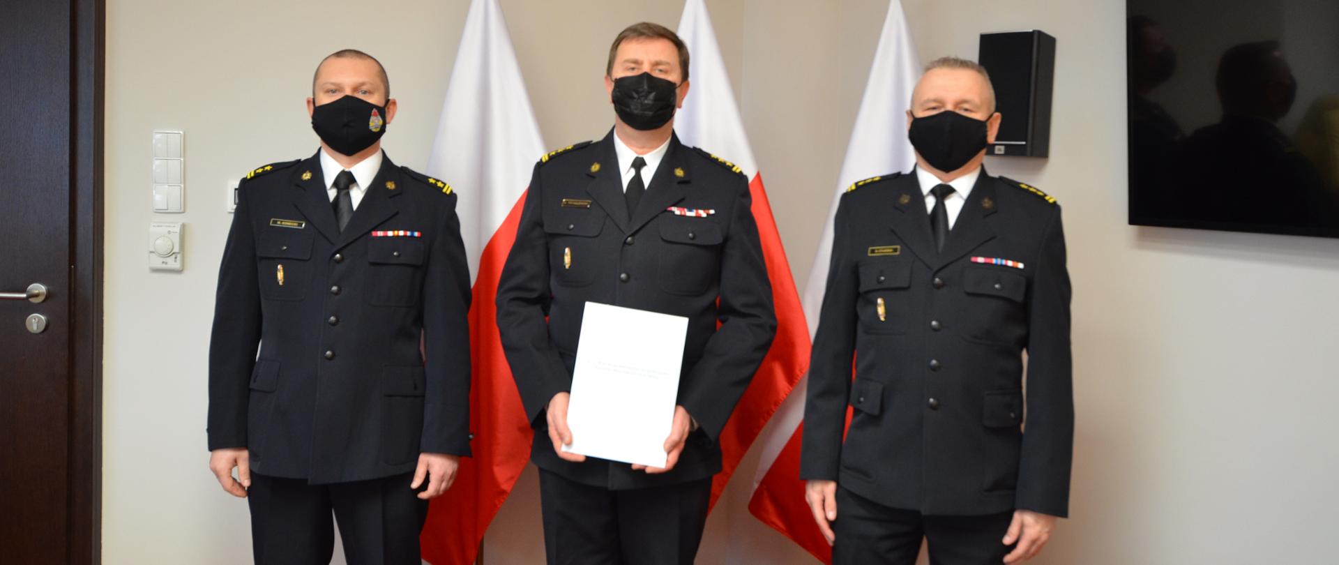 Na zdjęciu widać trzech strażaków w umundurowaniu wyjściowym. W tle trzy flagi Polski.