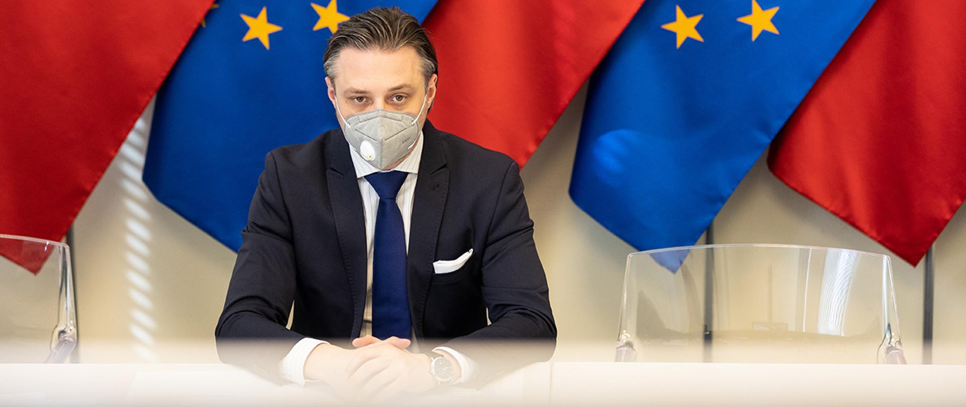 Na zdjęciu widać wiceministra Bartosza Grodeckiego siedzącego na tle flag Polski i UE.