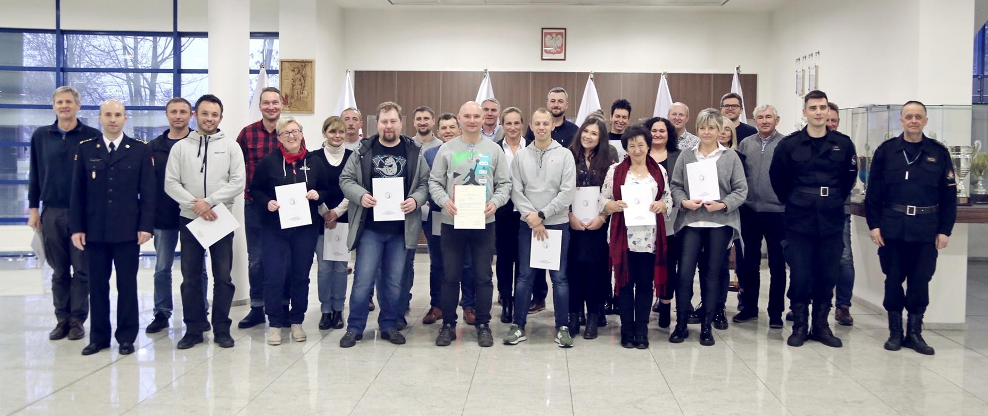 Grupowe zdjęcie uczestników szkolenia (26 osób) w towarzystwie Zastępcy Komendanta Centralnej Szkoły PSP, dwóch funkcjonariuszy CS PSP oraz kobiety. Uczestnicy szkolenia trzymają otrzymane zaświadczenia