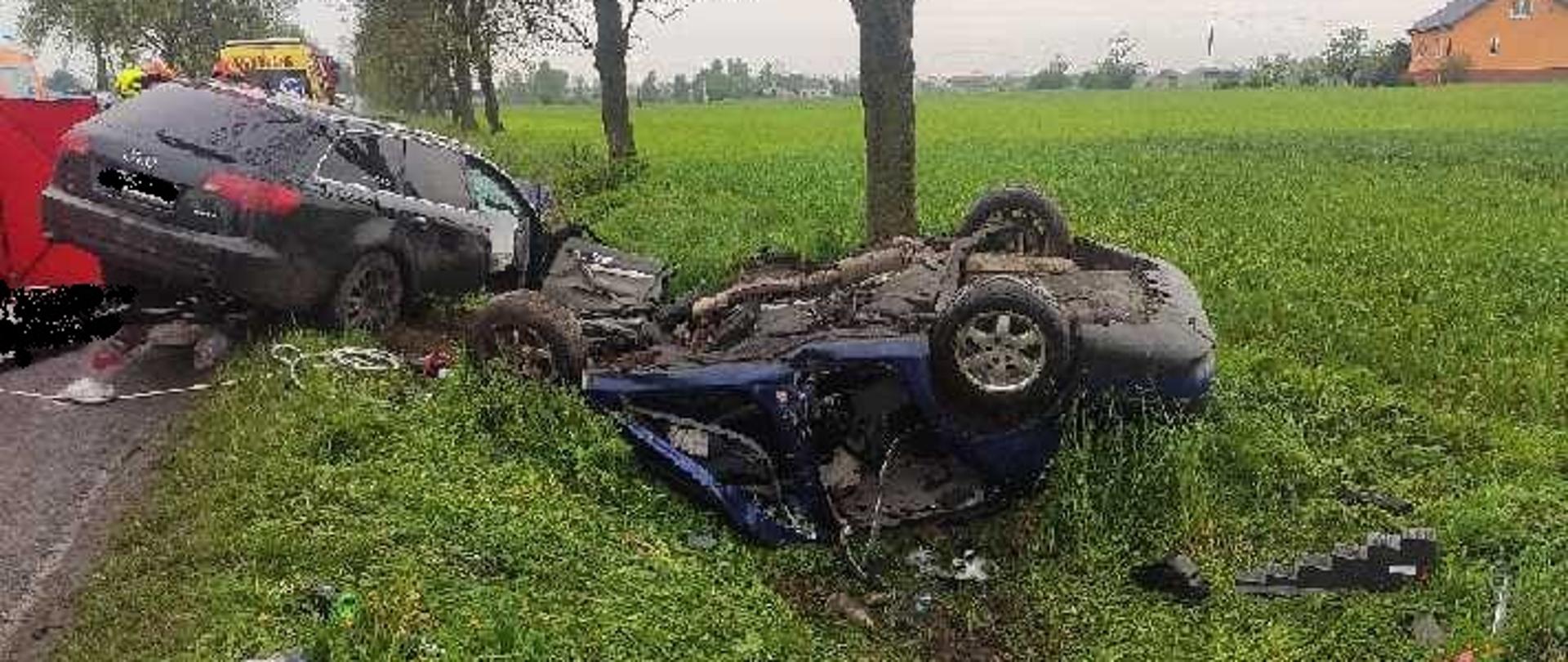 Zdjęcie przedstawia dwa samochody osobowe po zderzeniu. Czarny samochód marki Audi A6 częściowo znajduje się na pasie jezdni i na pobliskim poboczu. Drugi samochód - niebieski Seat Toledo znajduje się na dachu na poboczu jezdni. Przy pojazdach widać podpory oraz narzędzia hydrauliczne. Oba pojazdy są bardzo zniszczone. W tle zdjęcia widać ZRM i strażaków. 