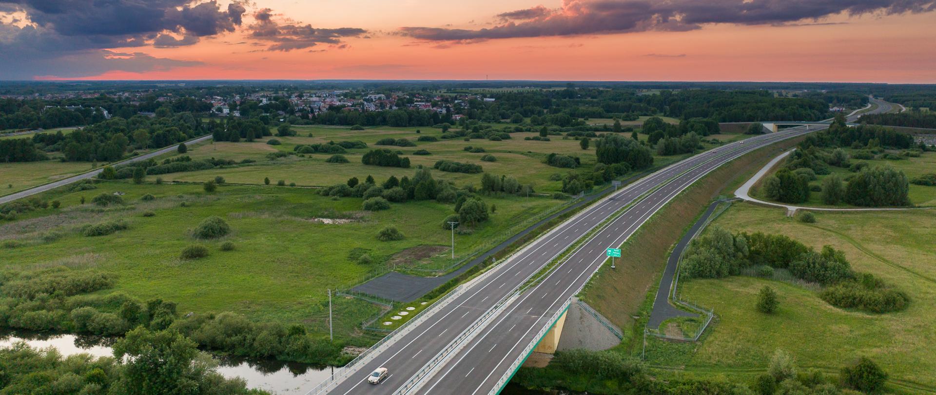 Widok z lotu ptaka na drogę ekspresową S19 przecinającą zielone pola i rzekę; w tle zabudowania i zachód słońca z kolorowymi chmurami.”
