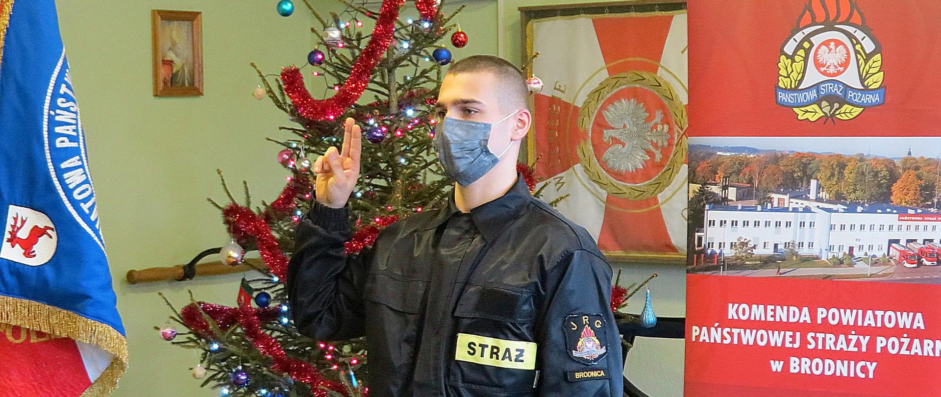 Zdjęcie przedstawia moment ślubowania - strażak Kacper Gmiński z uniesionymi palcami dłoni wypowiada rotę ślubowania. W tle sztandar Komendy oraz ustrojona choinka.