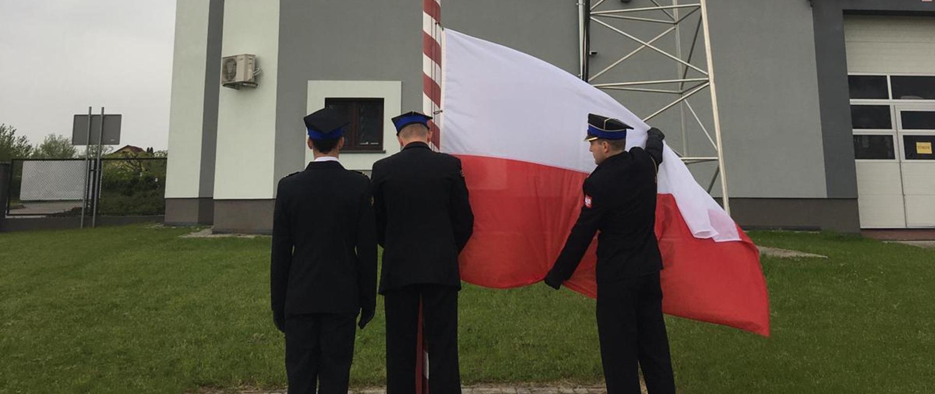 Troje strażaków w umundurowaniu wyjściowym zawiesza flagę Polski na maszt.