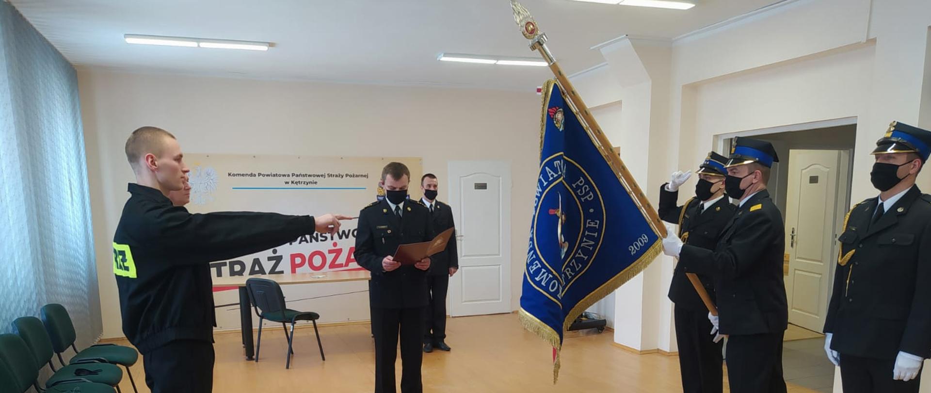 W sali konferencyjnej komendy nowoprzyjęty strażak unosi prawą rękę do sztandaru. W tle komendant powiatowy PSP, a po prawej trzech strażaków wchodzących w skład pocztu sztandarowego. Strażacy ubrani w czarny mundur wyjściowy, strażak składający ślubowanie w czarny mundur koszarowy.