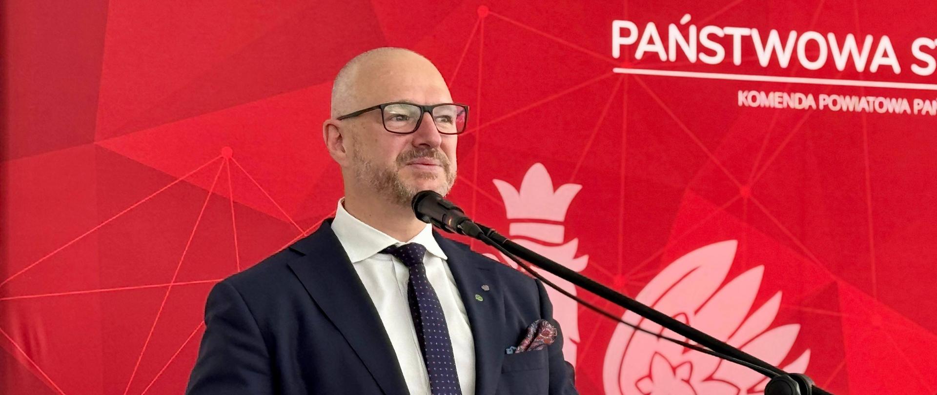 Mężczyzna przemawia na tle czerwonej ścianki z napisem Państwowa Straż Pożarna 