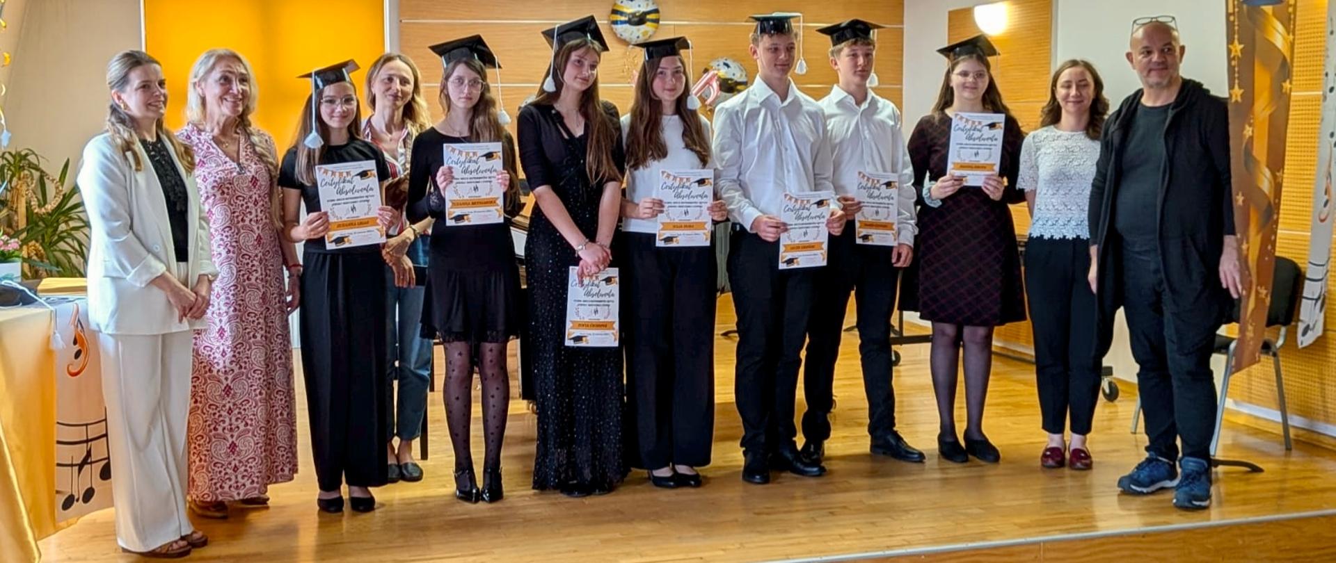 W centralnej części stoi siedmioro młodych ludzi (pięć dziewcząt i dwóch chłopców). Wszyscy mają na głowach czarne czapki studenckie (birety) z frędzlami i trzymają w dłoniach pamiątkowe dyplomy z napisem „Certyfikat Absolwenta”. Dziewczęta są ubrane w eleganckie, ciemne sukienki lub bluzki i spodnie, natomiast chłopcy mają na sobie klasyczne białe koszule i ciemne spodnie.Po lewej i prawej stronie grupy absolwentów stoją dorośli – łącznie pięć osób. Po lewej stronie widać m.in. kobietę w białym garniturze oraz kobietę w długiej, wzorzystej sukience. Po prawej stronie stoi młoda kobieta w jasnej, koronkowej bluzce oraz mężczyzna w ciemnym ubraniu z okularami opartymi na czubku głowy. Scena ma jasną, drewnianą podłogę. W tle znajduje się ściana z drewnianych paneli, żółta roleta okienna oraz dekoracyjne balony. Po prawej stronie widać fragment ozdobnego baneru z motywem biretów i gwiazd. Wszystkie osoby są zwrócone przodem do obiektywu i uśmiechają się.