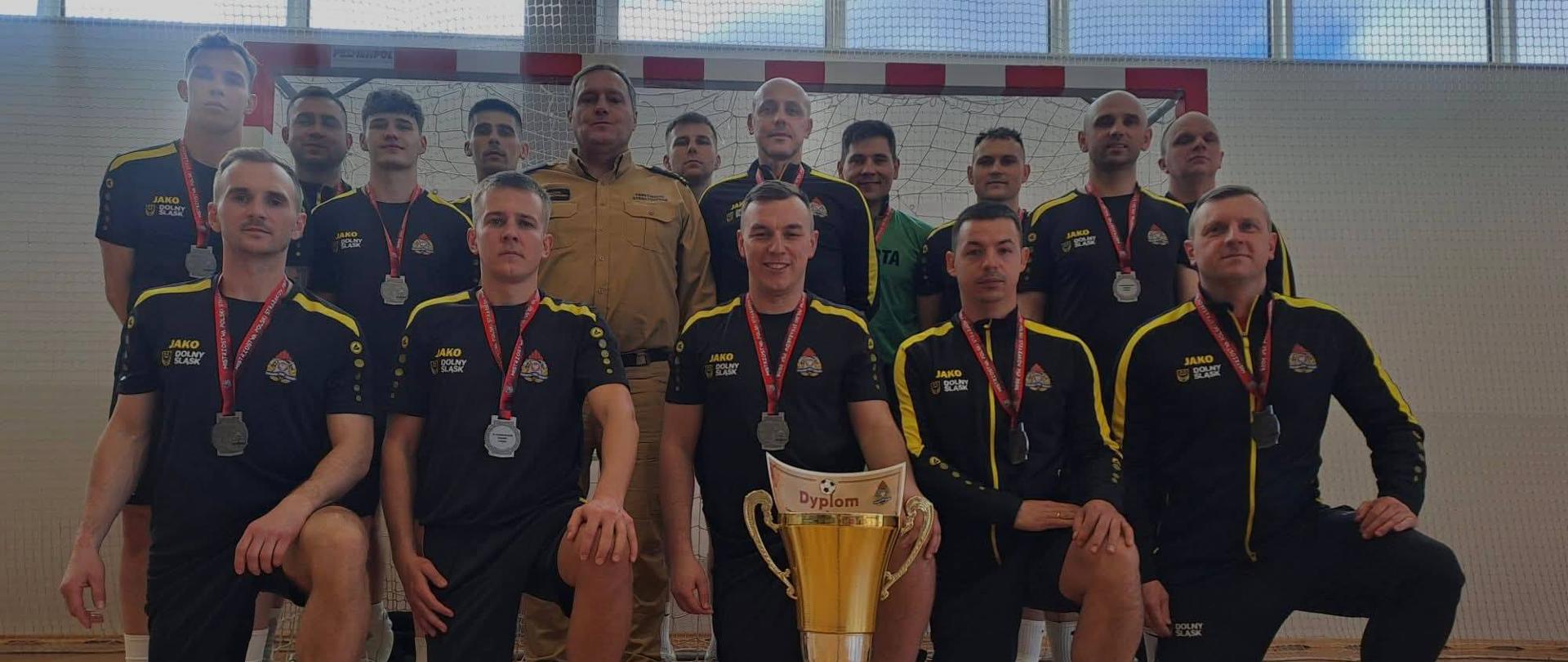 Zdjęcie przedstawia grupę mężczyzn, drużynę sportową, pozującą do pamiątkowej fotografii w hali sportowej po zakończonych zawodach.