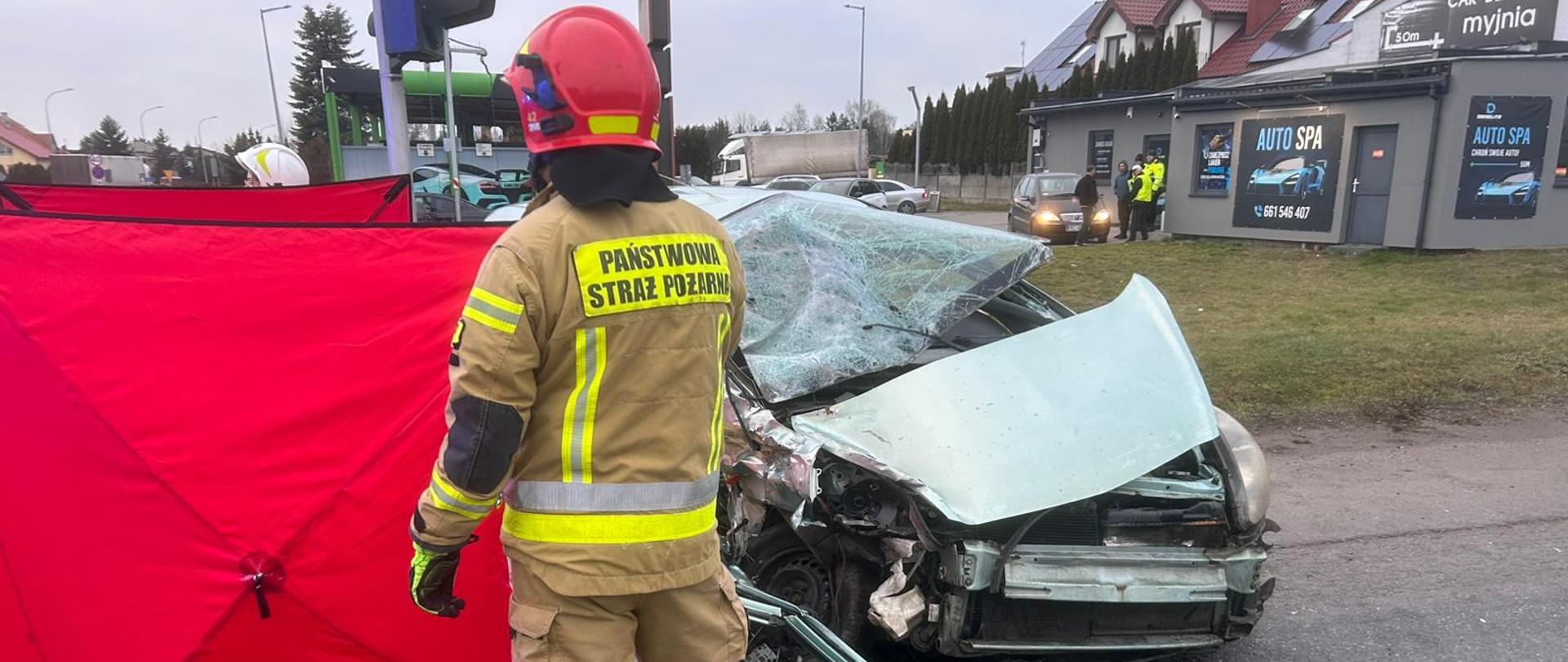 Na zdjęciu widać strażaka Państwowej Straży Pożarnej stojącego tyłem, ubranego w ubranie ochronne i czerwony hełm, obok poważnie uszkodzonego samochodu osobowego po zderzeniu na skrzyżowaniu. Przód auta jest mocno zgnieciony, maska i szyba czołowa są zniszczone. Obok pojazdu rozstawiony jest czerwony parawan osłaniający miejsce zdarzenia, a w tle widoczne są inne samochody, zabudowania oraz sygnalizacja świetlna.