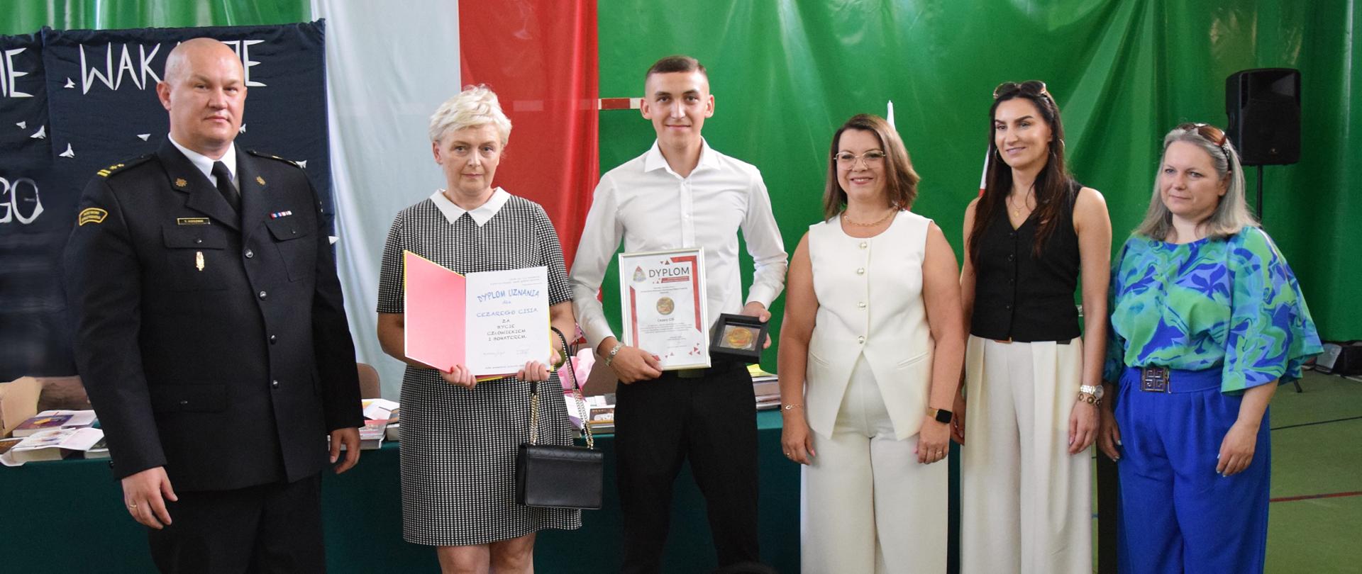 6 osób ubranych w eleganckie stroje stoi w szeregu na tle zielonej ściany z flagą polski. Za nimi stół w nagrodami i książkami. Wszyscy się uśmiechają, Młody uczeń stojący po środku trzyma w rękach dyplom oraz Odznakę, kobieta po jego lewej stronie na zdjęciu trzyma otwarty dyplom.