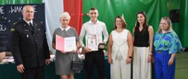 6 osób ubranych w eleganckie stroje stoi w szeregu na tle zielonej ściany z flagą polski. Za nimi stół w nagrodami i książkami. Wszyscy się uśmiechają, Młody uczeń stojący po środku trzyma w rękach dyplom oraz Odznakę, kobieta po jego lewej stronie na zdjęciu trzyma otwarty dyplom.