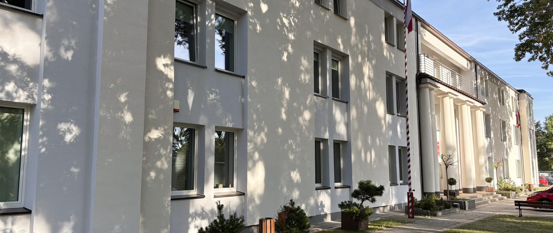 Budynek Ośrodka Szkolenia w Pionkach Komendy Wojewódzkiej Państwowej Straży Pożarnej w Warszawie po modernizacji. Dwupiętrowy obiekt z jasną elewacją, otoczony drzewami i chodnikami, widoczny w słoneczny dzień.
