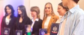 Na scenie stoi sześcioro absolwentów trzymających w rękach świadectwa ukończenia szkoły w teczkach z orłem i biało-czerwoną flagą