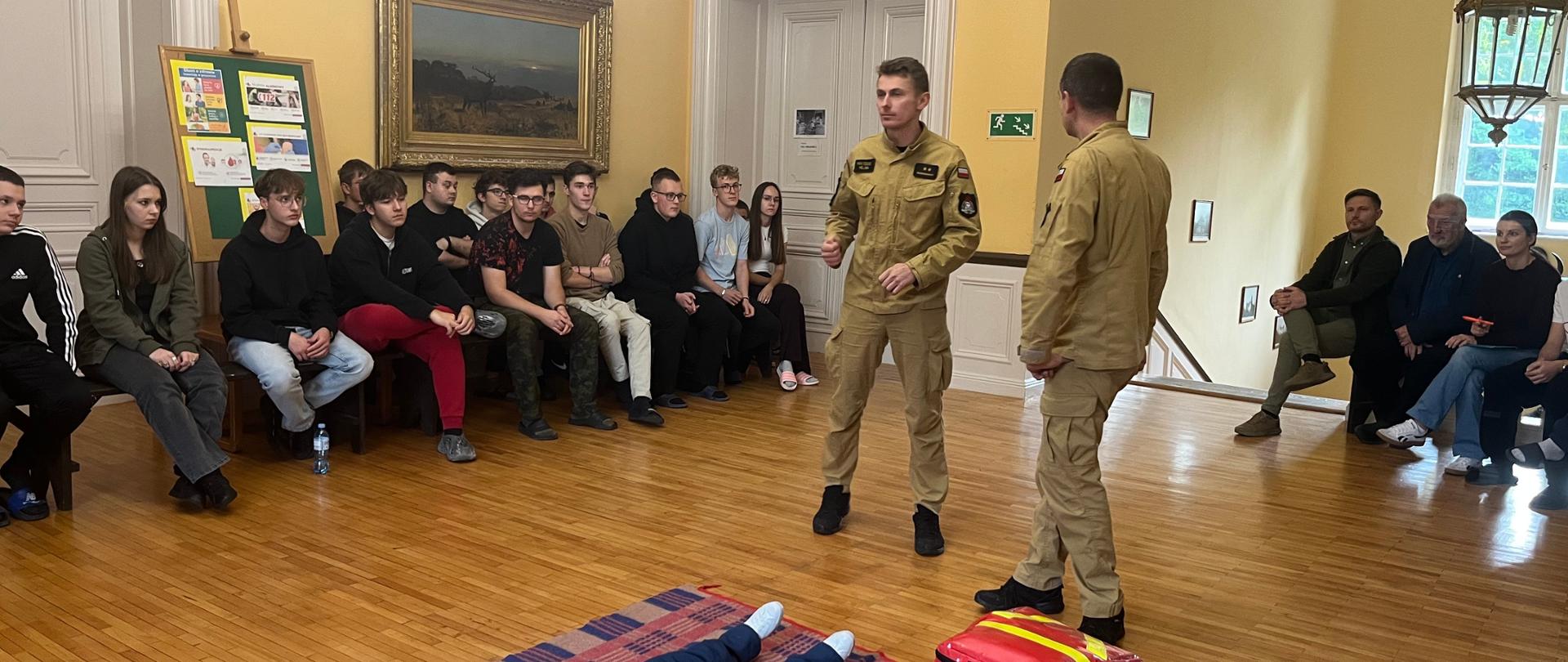 Strażacy z JRG PSP Czarnków podczas pokazu udzielania pierwszej pomocy – w sali szkoły zebrani uczniowie obserwują instruktaż przy fantomie.