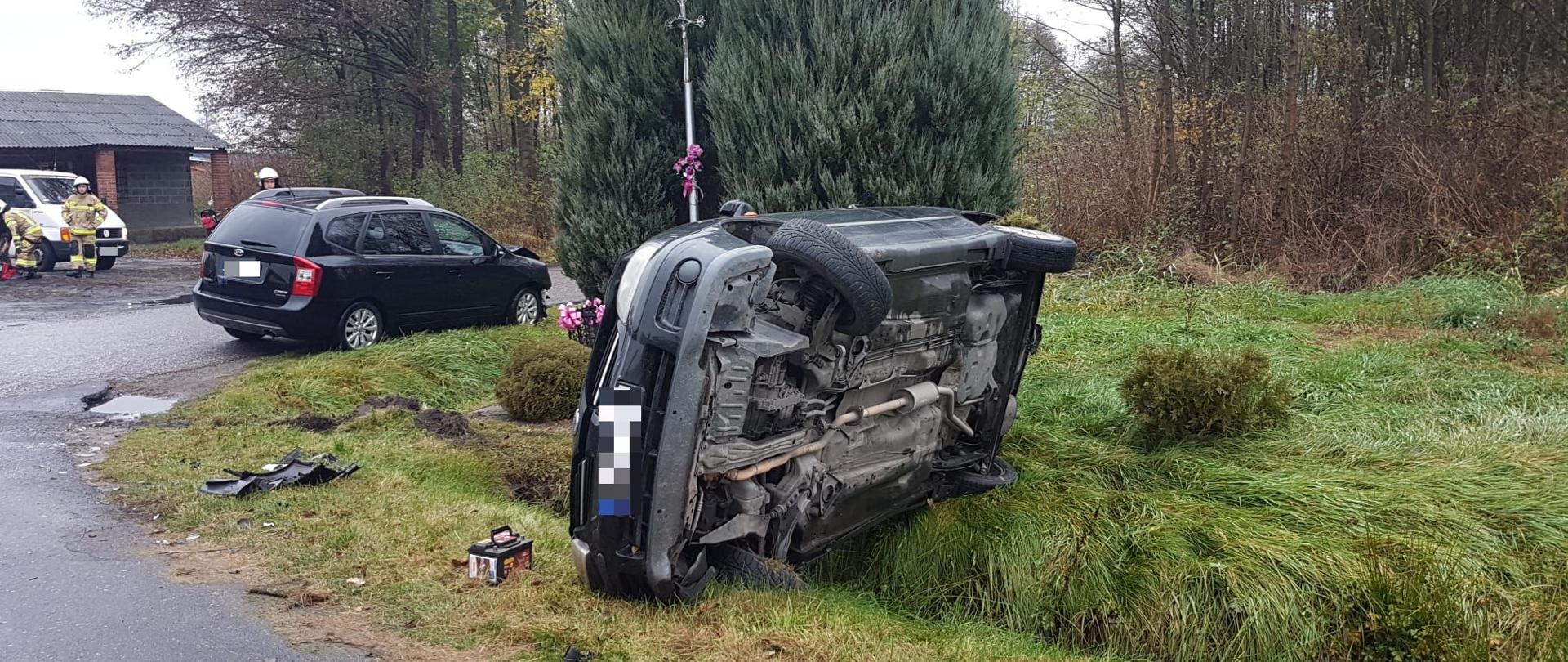 na zdjęciu widać dwa samochody koloru czarnego, które brały udział w zdarzeniu jeden lezy na boku drugi stoi na poboczu za samochodem który lezy na boku widać krzyż oraz dwa drzewa tuje koloru zielonego w pobliżu porozbijane elementy samochodu. w tle trzech strażaków obok białego samochodu dalej czerwony budynek gospodarczy oraz drzewa.