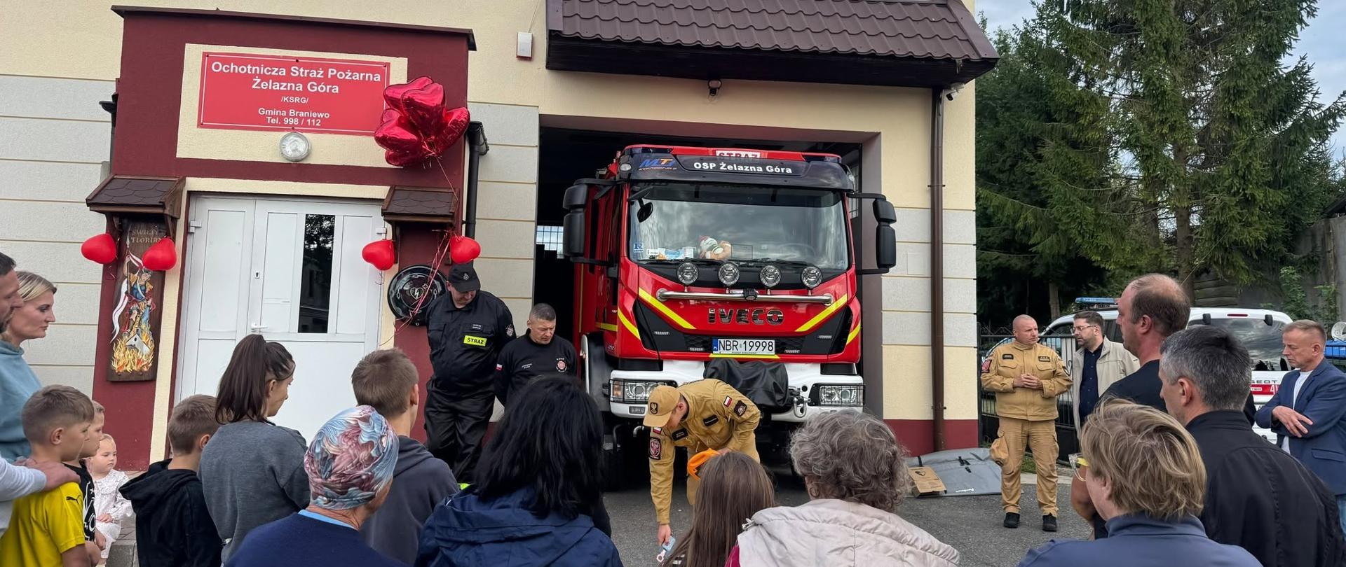 Przed remizą Ochotniczej Straży Pożarnej w Żelaznej Górze zgromadzili się mieszkańcy, w tym dzieci, oraz strażacy. W otwartym garażu stoi czerwony samochód ratowniczo-gaśniczy marki Iveco. Jeden ze strażaków prezentuje sprzęt rozłożony przed pojazdem, a zgromadzeni obserwują pokaz. Budynek remizy ozdobiono czerwonymi balonami, co nadaje spotkaniu uroczysty charakter.