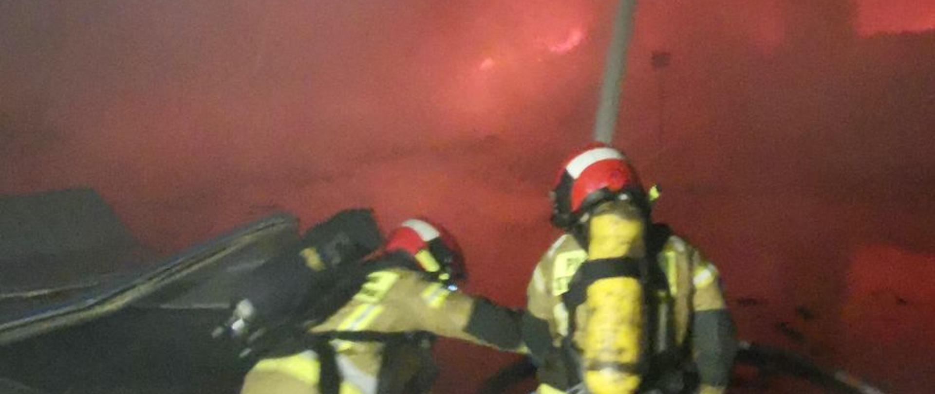 Zdjęcie przedstawia dwóch strażaków gaszących pożar we wnętrzu hali produkcyjnej