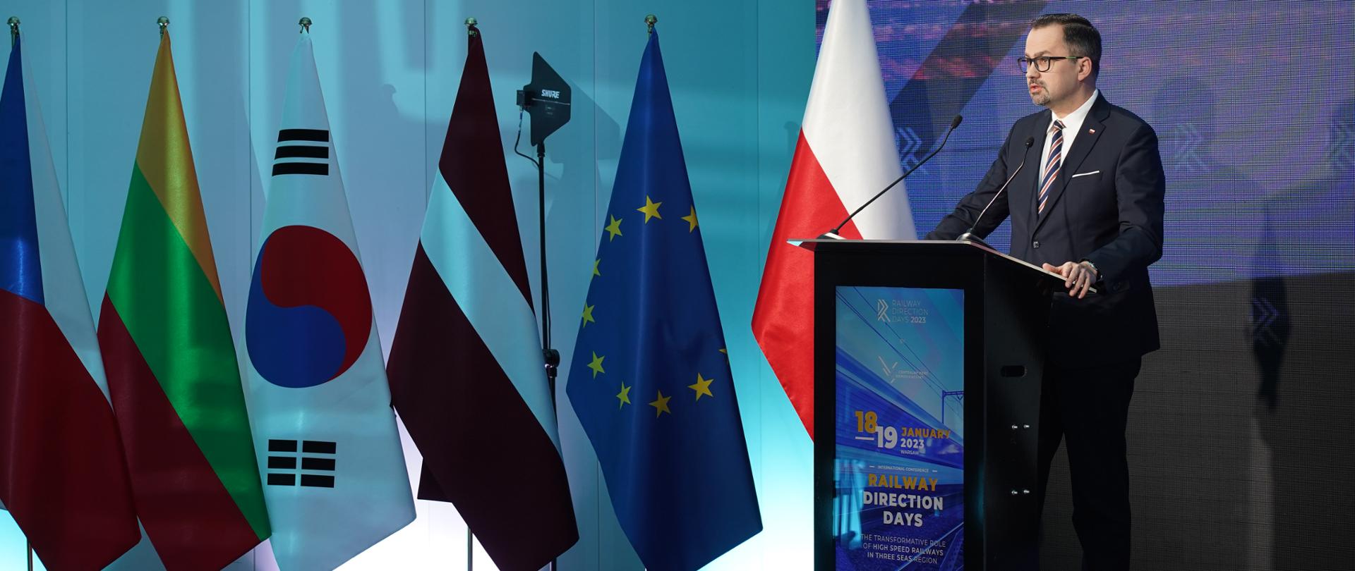 Wiceminister Marcin Horała stoi na scenie przy podeście i mówi do mikrofonu.