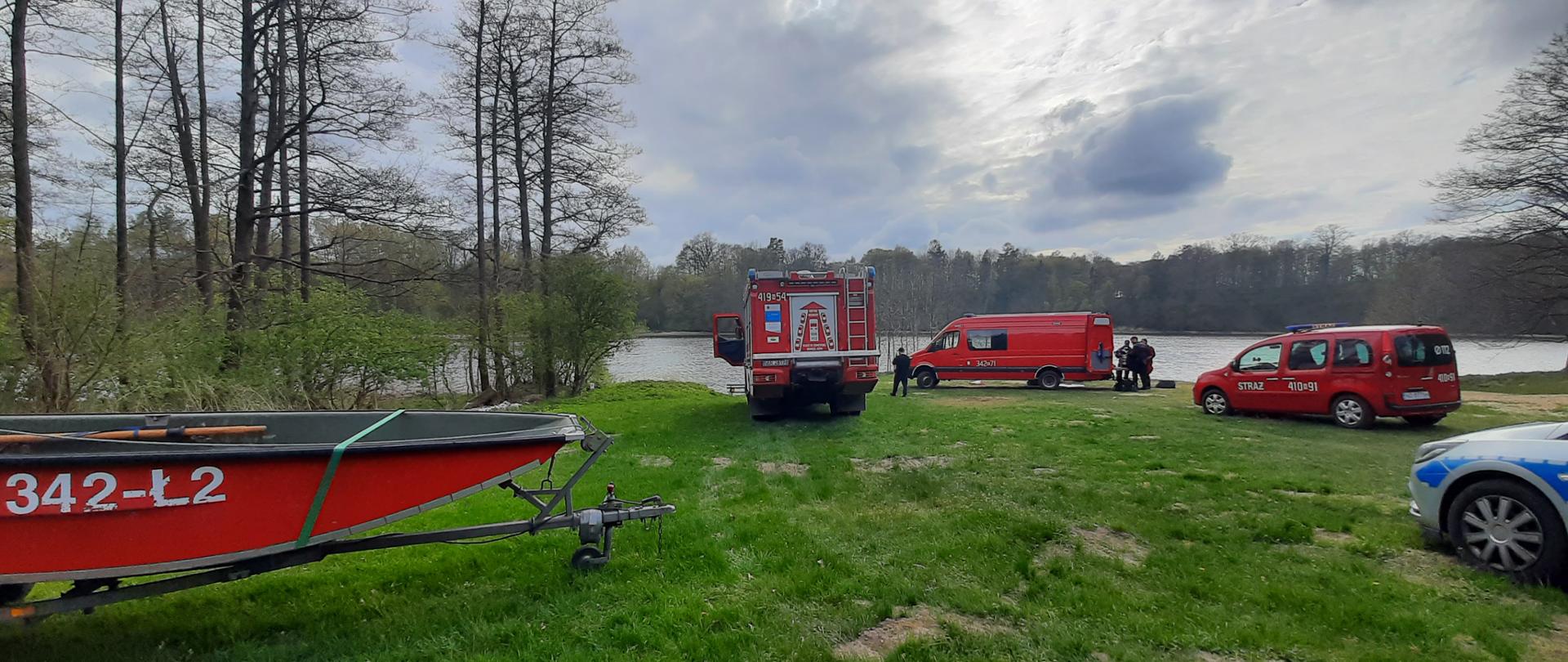 Zielona polana nad jeziorem. Z lewej strony stoi czerwona lódź strażacka a z prawej i centralnie trzy wozy strażackie. W oddali jezioro i las. Pochmurnie.