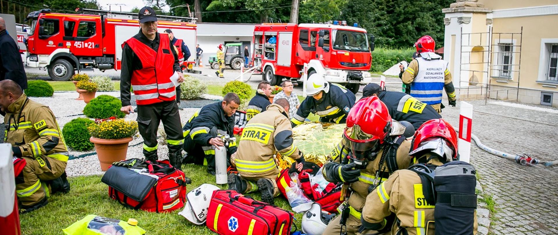 Strażacy udzielający pomocy pozorantom uczestniczącym w Ćwiczeniach Powiatowych, Ratowniczy pochyleni nad poszkodowanymi znajdującymi się na deskach ratowniczych. W czerwonych kamizelkach nad ratownikami stoją oceniający wykonywane czynności mediatorzy. W tle zdjęcia dostrzegamy ustawione bokiem dwa pojazdy ratowniczo gaśnicze w kolorze czerwonym z widocznymi kryptonimami.
