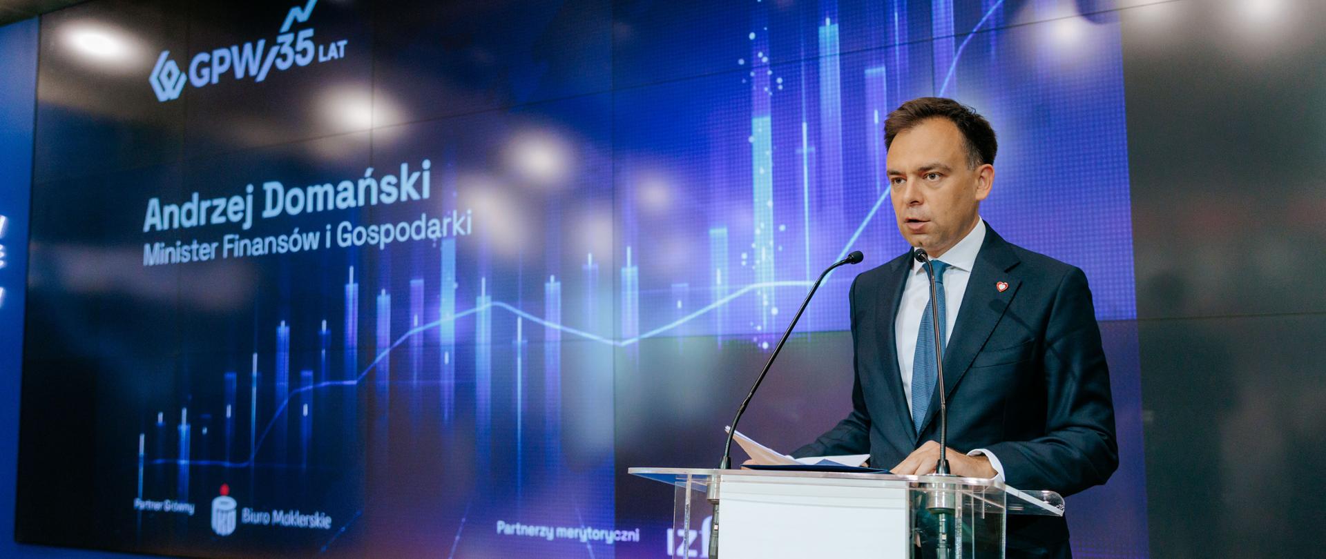 Minister finansów i gospodarki Andrzej Domański podczas wystąpienia na Giełdzie Papierów Wartościowych na tle telebimu, za mównicą. Źródło zdjęcia: GPW