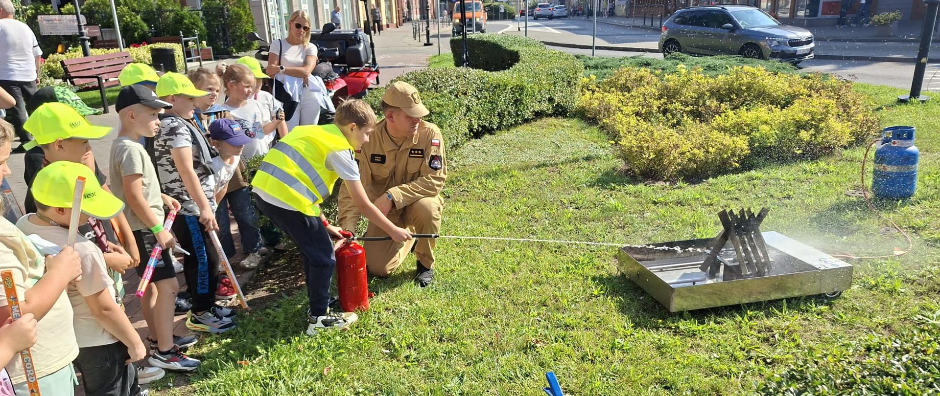 Strażak pomaga dzieciom w obsłudze gaśnicy podczas ćwiczeń na świeżym powietrzu.