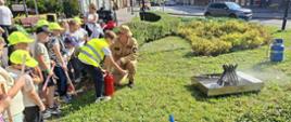 Strażak pomaga dzieciom w obsłudze gaśnicy podczas ćwiczeń na świeżym powietrzu.