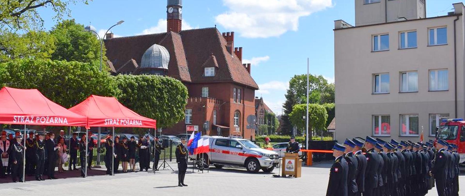 Plac KP PSP Tczew. Po prawej stronie stoi pododdział w umundurowaniu wyjściowym frontem do dwóch namiotów, pod którymi stoją zaproszeni goście, przy mównicy przemawia Komendant, w tle ratusz tczewski.