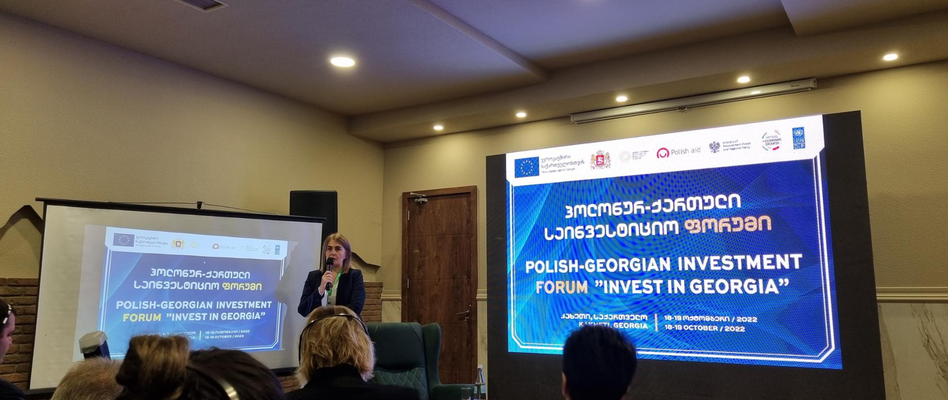 Kobieta przy mikrofonie stoi obok dwóch ekranów, na którym wyświetlają się napisy w języku angielskim i gruzińskim.