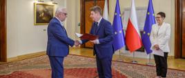 Minister Jan Grabiec wręcza akt powołania do Rady KSAP prof. Jerzemu Stępniowi, obok stoi szefowa służby cywilnej Anita Noskowska-Piątkowska; w tle flagi Polski i Unii Europejskiej