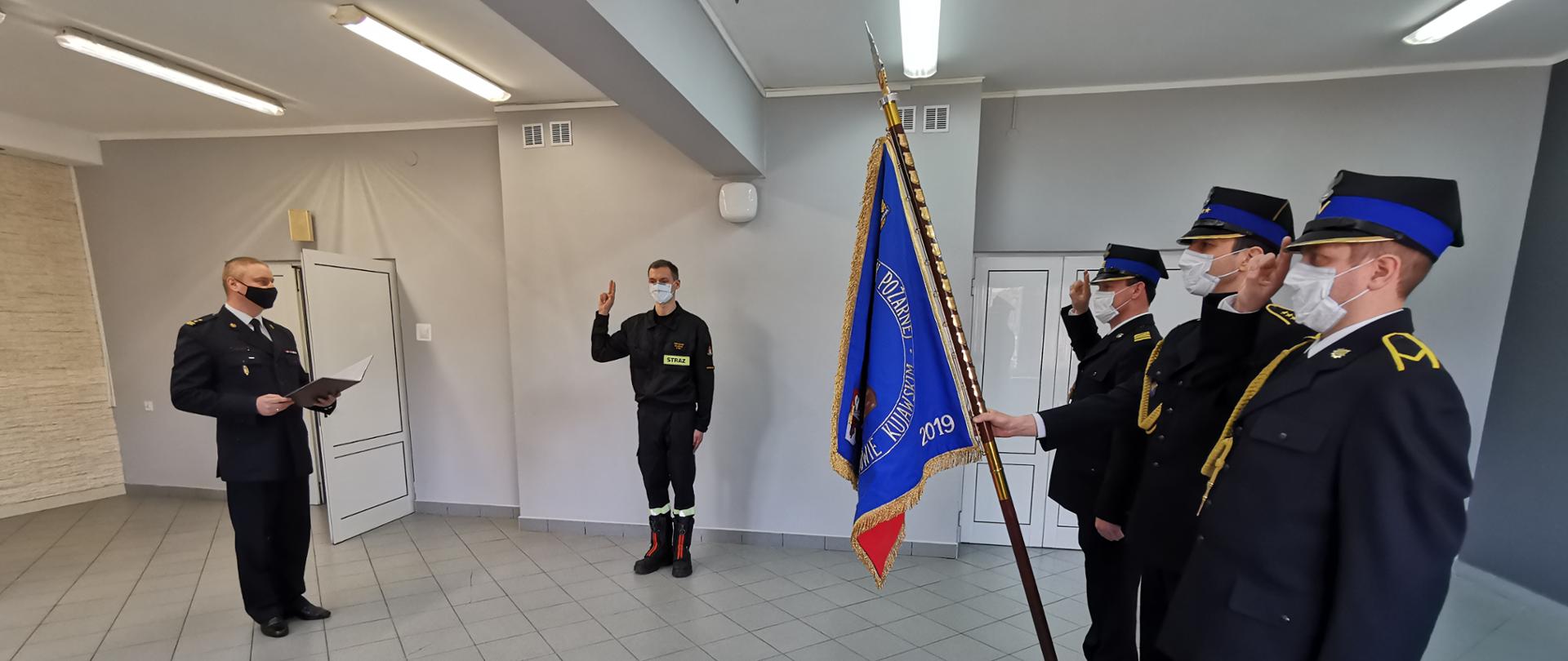 Zdjęcie obrazuje ślubowanie nowoprzyjętego strażaka do służby w PSP. Z prawej strony zdjęcia widoczni są strażacy z pocztu sztandarowego w umundurowaniu wyjściowym. Z lewej strony widoczny Komendant Powiatowy, który odczytywał tekst Roty ślubowania. W centralne części zdjęcia widoczny strażak w umundurowaniu koszarowym.