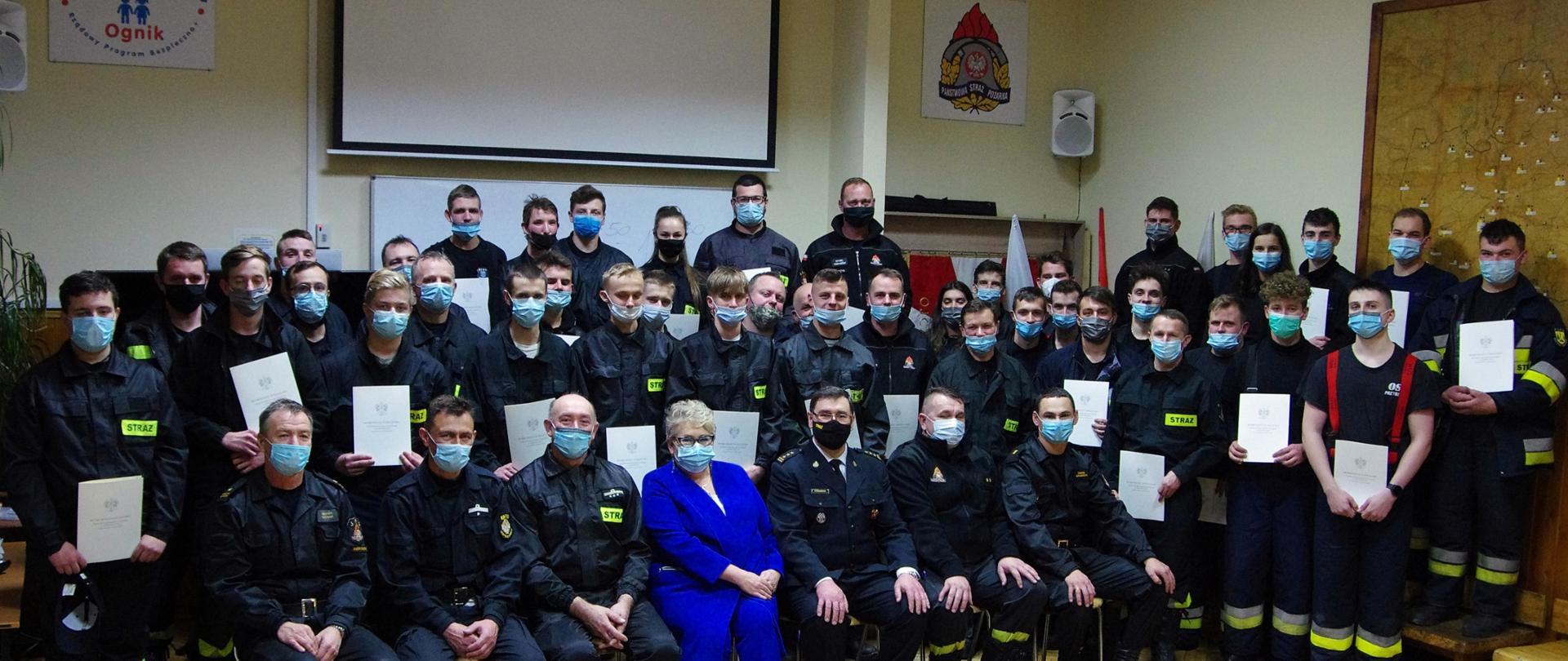 Widok z przodu. Zdjęcie zbiorowe wszystkich uczestników szkolenia. Wśród absolwentów szkolenia siedzi wicestarosta wadowicka w niebieskim ubraniu cywilnym oraz strażacy PSP i OSP w umundurowaniu. Wszystkie osoby w maseczkach ochronnych. 