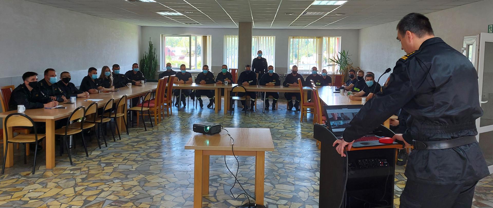 Na zdjęciu widać uczestników warsztatów medycznych. Na pierwszym planie Komendant Powiatowy, w oddali za stołami siedzą strażacy PSP i OSP. 