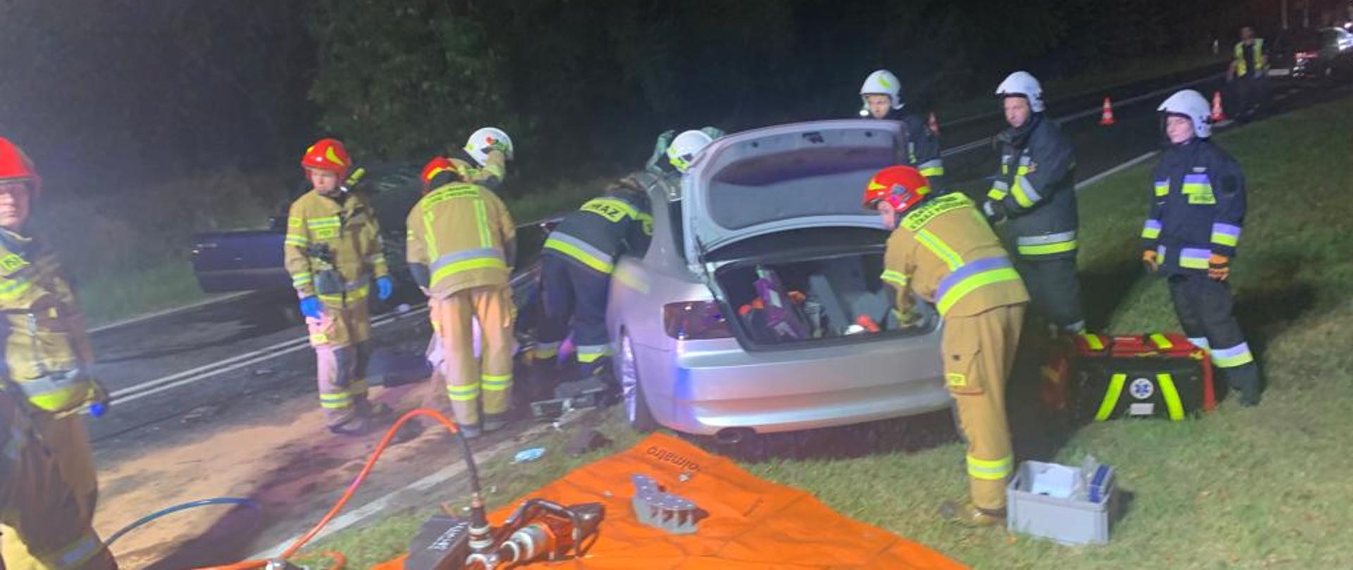 zdjęcie przedstawia samochód osobowy koloru srebrnego z podniesiona klapą bagażnika obok niego strażacy w ubraniach specjalnych przed samochodem pomarańczowa mata na której rozłożony jest sprzęt hydrauliczny 