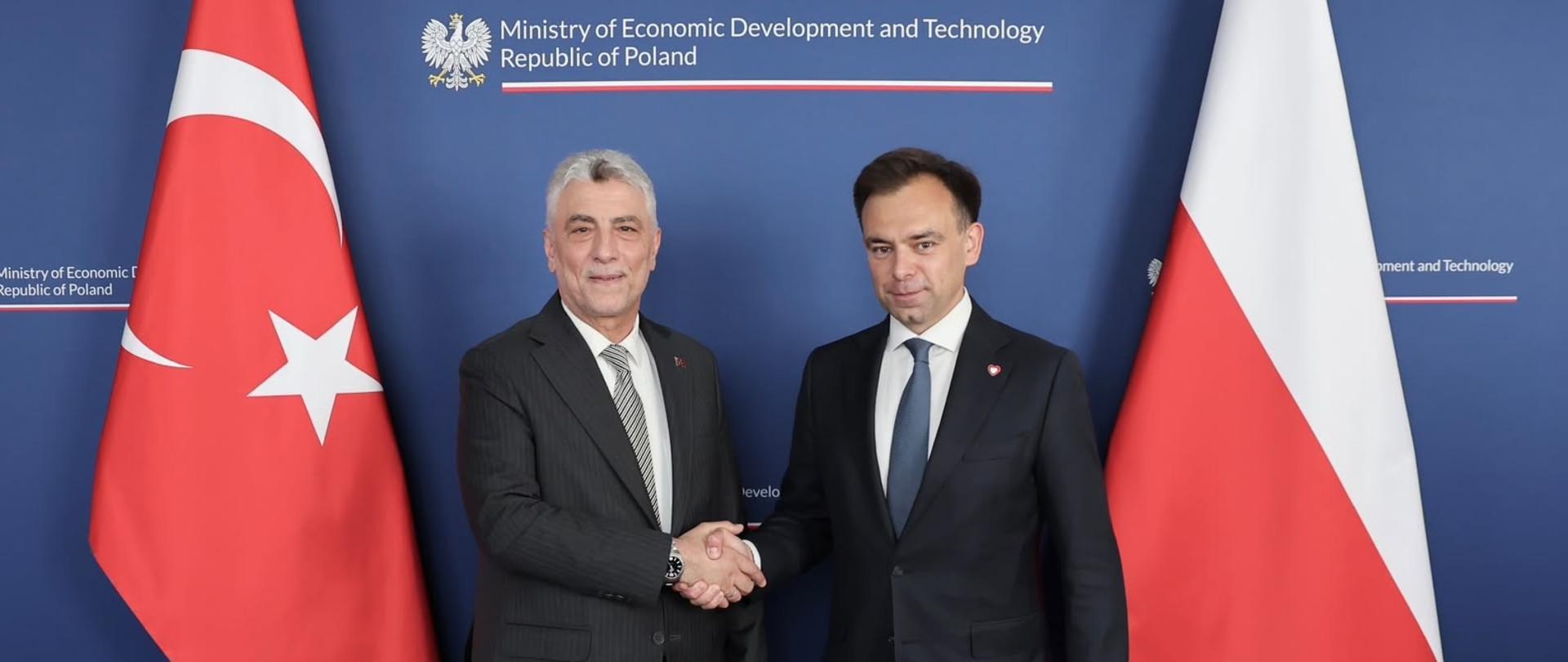 Oficjalny uścisk dłoni min. Andrzeja Domańskiego (z prawej) i ministra handlu Republiki Turcji Ömera Bolata (z lewej) na tle granatowej ścianki z logo MRIT. Przy ministrach flagi obydwu krajów.