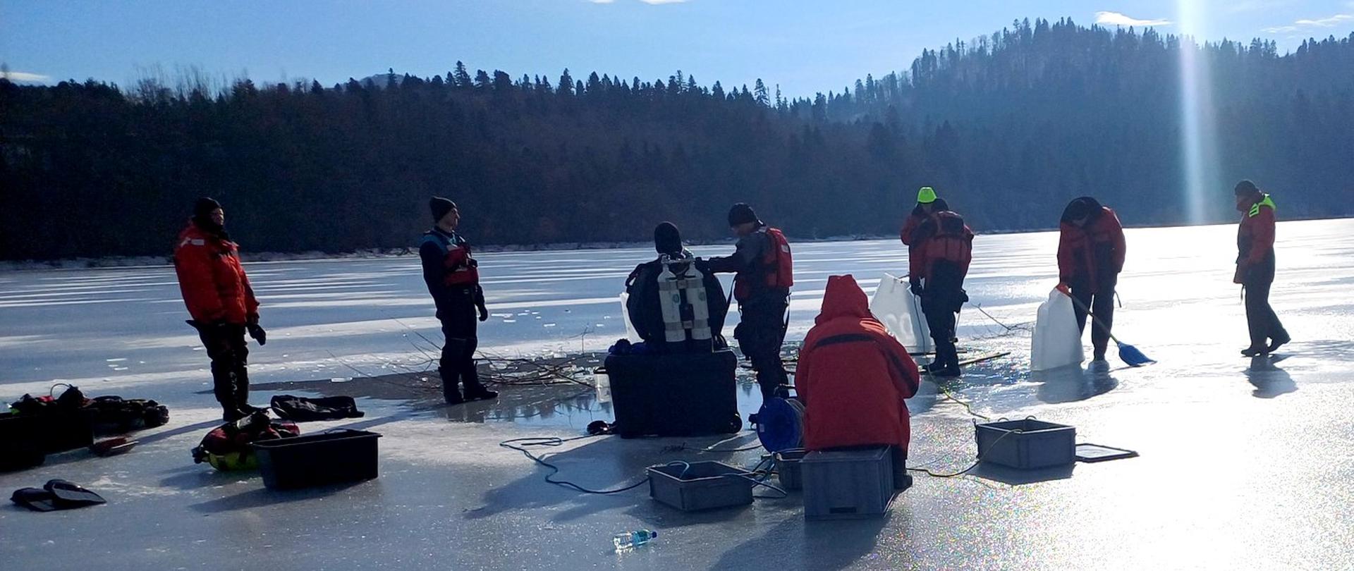 Na zdjęciu widać zalodzony zbiornik wodny w którym wycięty przerębel. W środku w wodzie nurek , na krawędzi przerębla siedzi tyłem nurek w sprzęcie do nurkowania. Nurków zabezpieczają koledzy strażacy ubrani w kamizelki ratunkowe i skafandry nurkowe. Trzymają liny. W oddali widać nadzorującego kierownika szkolenia w kamizelce do ratownictwa wodnego