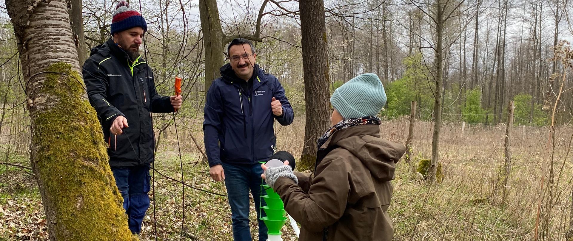 Grupa trzech osób prowadzi prace badawcze lub instalacyjne w naturalnym środowisku leśnym. Dwóch mężczyzn stoi przy drzewie, a trzecia osoba, ubrana w brązową odzież i czapkę, trzyma element sprzętu i rozmawia z nimi. Jeden z mężczyzn uśmiecha się i gestykuluje, podczas gdy drugi skupia się na przewodzie i narzędziu w dłoni. W tle rozciąga się otwarta przestrzeń z suchą roślinnością i młodymi drzewami.