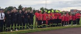 Zdjęcie przedstawia dużą grupę dorosłych i młodych ludzi stojących w rzędzie na trawiastym terenie, podczas zawodów strażackich. Dorośli po lewej stronie ubrani są w ciemne mundury strażackie, a po prawej stronie młodsi uczestnicy noszą czerwone koszulki i czarne spodnie, a niektórzy z nich mają na głowach jaskrawożółte kaski. Teren, na którym stoją, to bieżnia lub pas startowy, a za nimi widać zieleń drzew i zabudowania. Niebo jest pochmurne. 