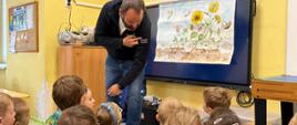 Inspektor prowadzi zajęcia edukacyjne dla grupy dzieci siedzących na podłodze w sali lekcyjnej. Obok niego znajduje się ekran z ilustracją przedstawiającą słoneczniki oraz owady i etapy ich rozwoju