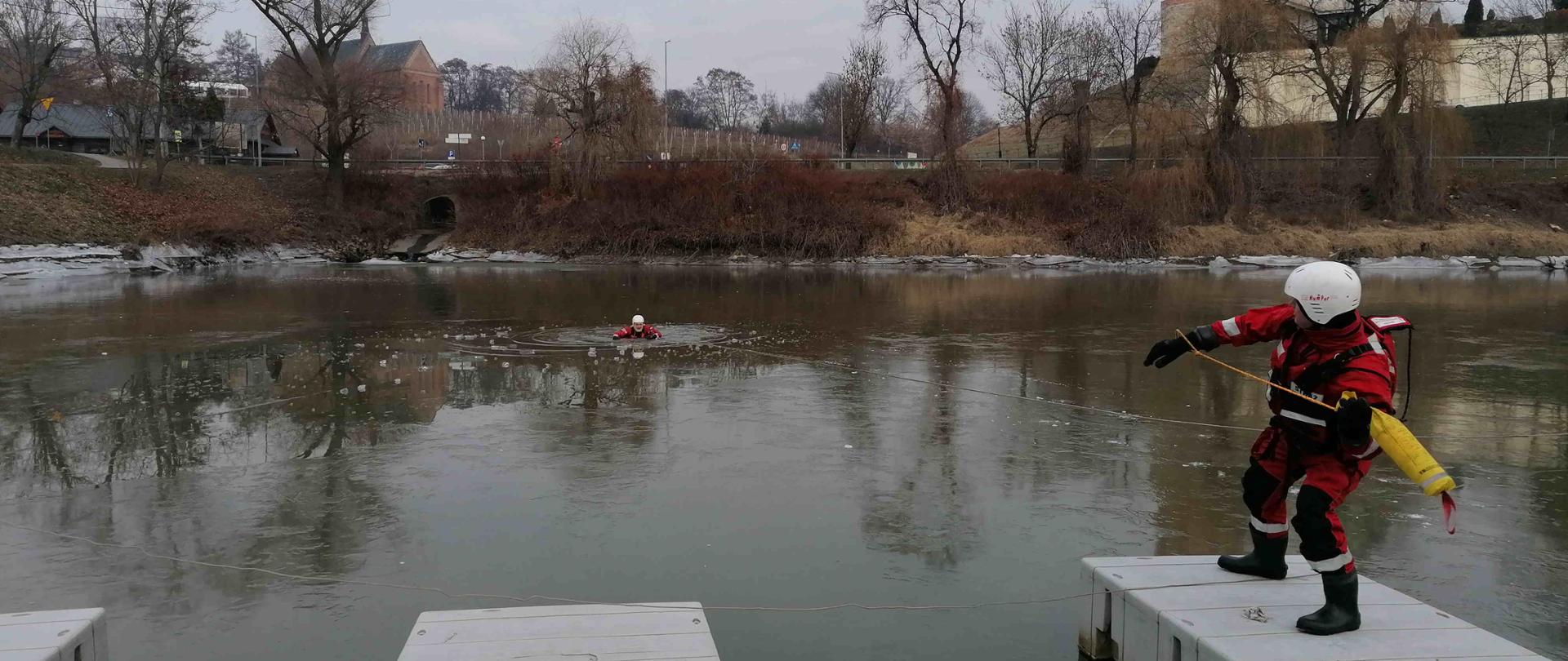 zdjęcie przedstawia zamarznięty zbiornik wodny i dwóch strażaków, którzy przy pomocy sań lodowych wyciągają poszkodowanego strażaka z wody.