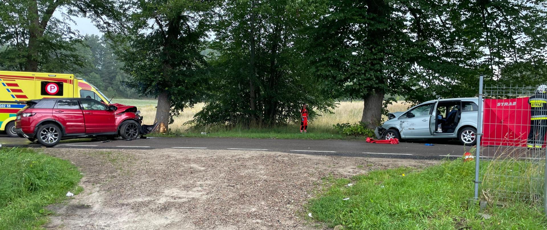 Po lewej stronie czerwony samochód osobowy, a za nim karetka pogotowia. Po prawej stronie srebrny samochód osobowy. 