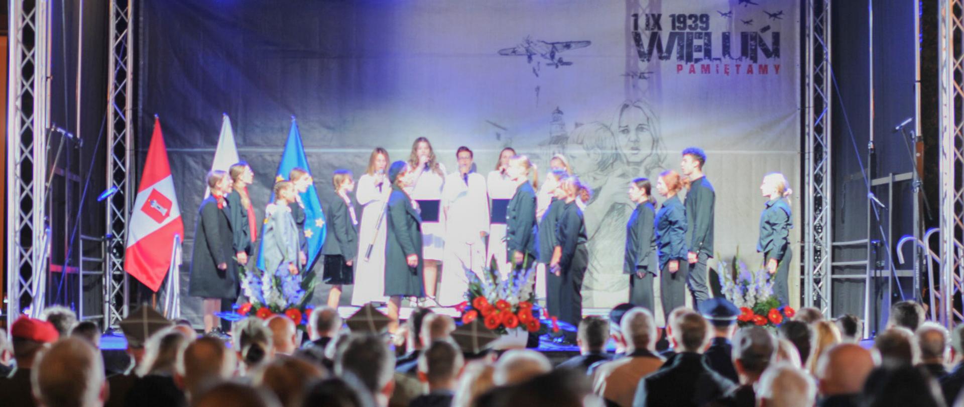 występ młodzieży na scenie z okazji upamiętnienie 84. rocznicy wybuchu II Wojny Światowej, przed sceną niewyraźni ludzie 