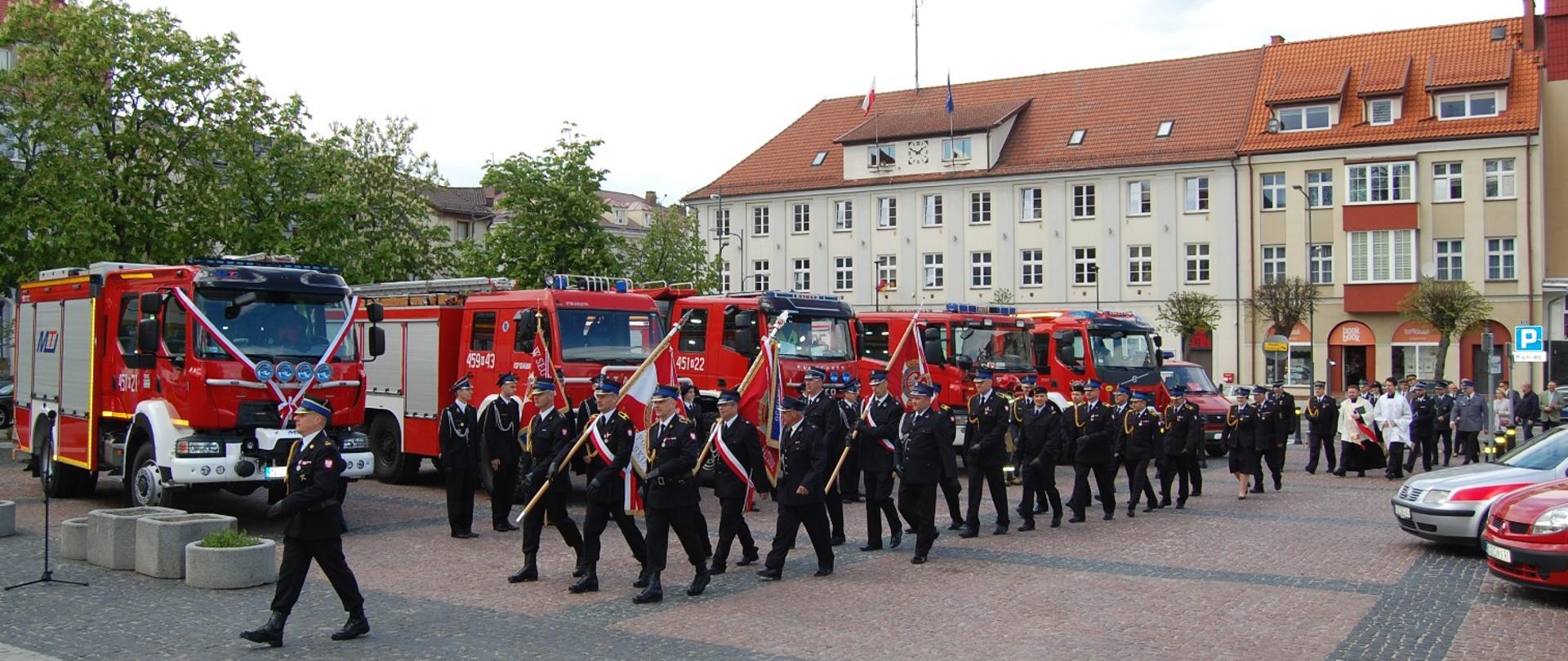 Zdjęcie przedstawia przemarsz pododdziału strażackiego oraz uczestników Mszy Świętej w intencji strażaków i ich rodzin z Kościoła pw. św. Leona Wielkiego w Gołdapi na Plac Zwycięstwa przy Pomniku Niepodległości w Gołdapi w celu kontynuowania obchodów Dnia Strażaka w formie uroczystego apelu. W tle budynki m.in. Urzędu Miejskiego w Gołdapi oraz pojazdy pożarnicze. 