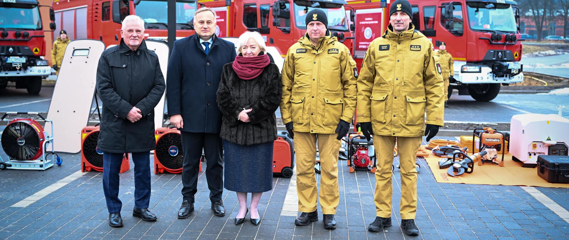 Grupa ludzi. z Tyłu sprzęt i samochody pożarnicze.