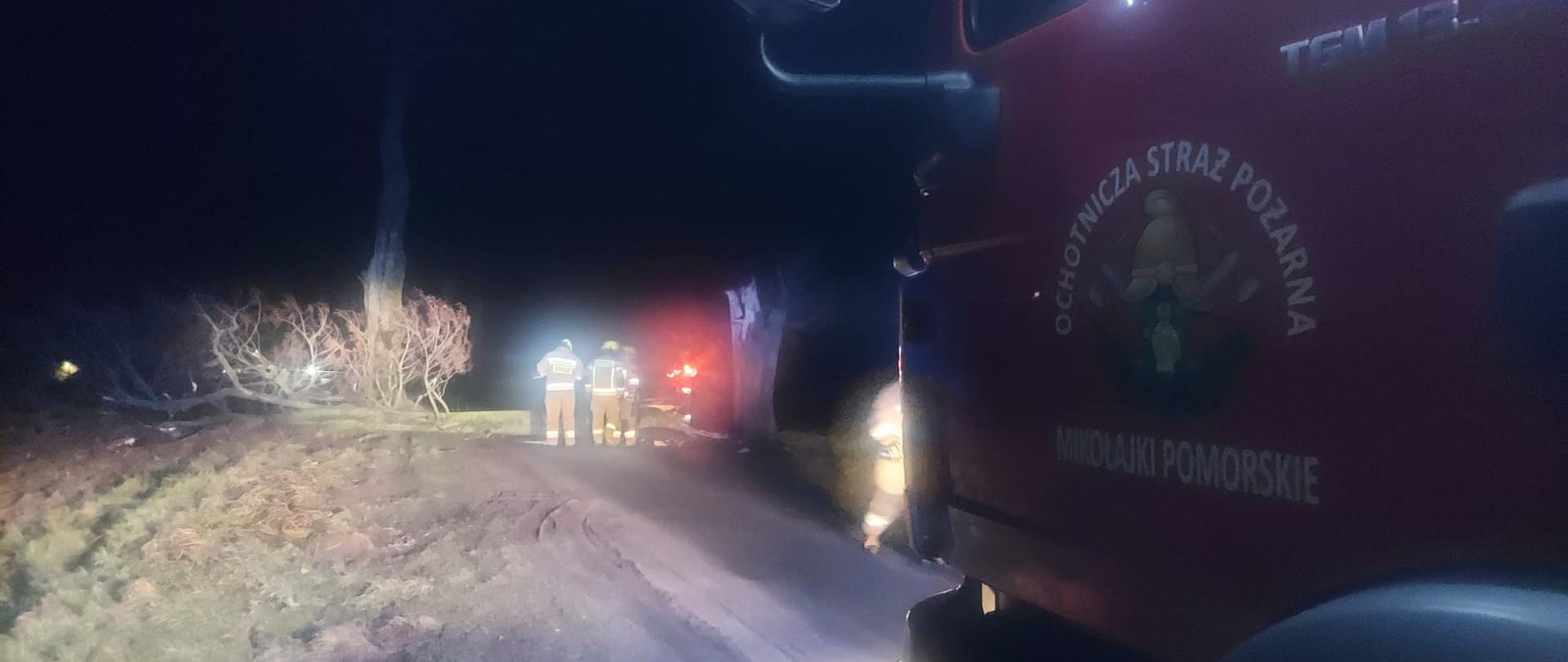 Pora nocna. na drodze stoi po prawej stronie (widoczny tylko przód) samochód pożarniczy. W oddali przed nim ratownicy usuwają powalone drzewo.