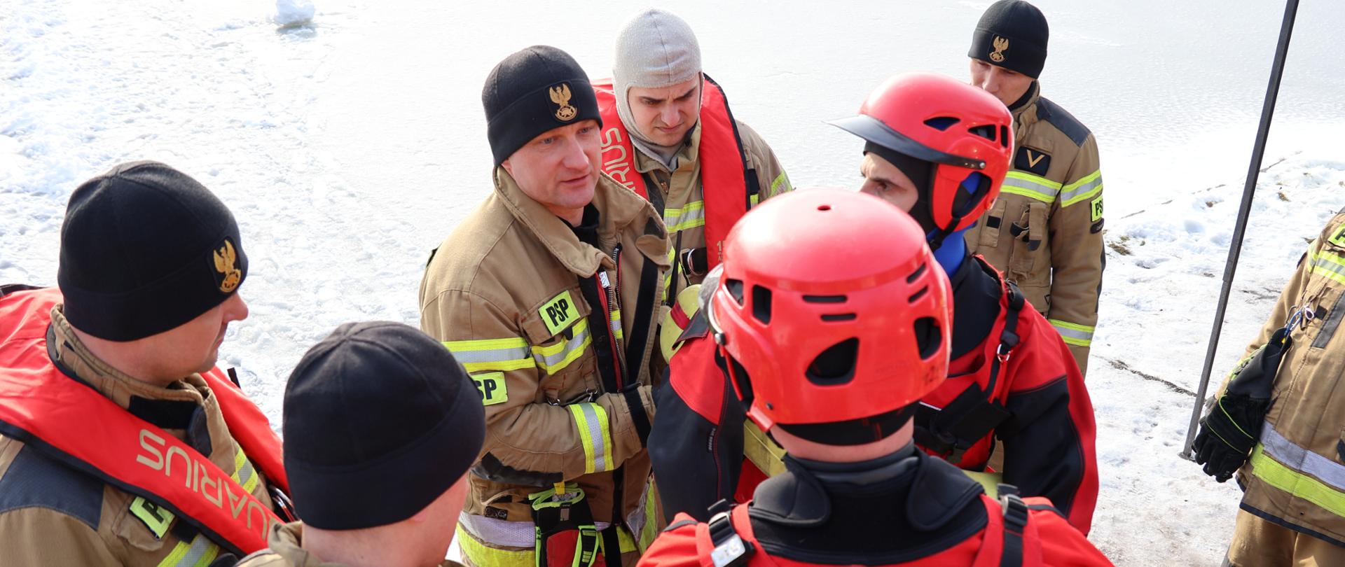 
Grupa ratowników z Państwowej Straży Pożarnej uczestniczy w zimowych ćwiczeniach ratowniczych na zamarzniętym akwenie. Ubrani w specjalistyczne kombinezony, kamizelki asekuracyjne i hełmy ochronne, strażacy prowadzą odprawę operacyjną na pokrytym śniegiem lodzie. Widoczne oznaczenia „PSP” i „STRAŻ” podkreślają ich przynależność służbową. Sceneria wskazuje na realistyczne warunki terenowe, a obecność sprzętu ratowniczego sugeruje przygotowanie do symulacji akcji ratunkowej z udziałem osób pod którymi załamał się lód. Ćwiczenia mają na celu doskonalenie współpracy zespołowej, komunikacji oraz skutecznego reagowania w sytuacjach zagrożenia życia w warunkach zimowych.