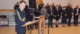 Wręczenie promes. Sala sportowa w Szkaradowie. Na pierwszym planie, stojący przy mównicy komendant powiatowy PSP w Rawiczu, który przemawia do zebranych. Na przodzie mównicy logotyp PSP. W tle gości uroczystej zbiórki - przedstawiciele władz państwowych i samorządowych oraz strażacy OSP