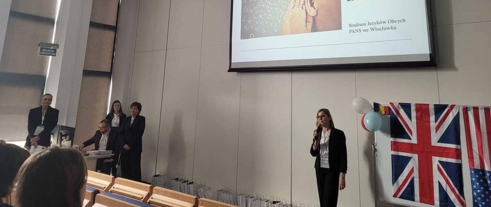 Na zdjęciu widać 4 uczniów i jednego nauczyciela, stojacego pomiedzy nimi. Osoby są na tle białe, wysokiej j sciany. Za nimi widać flagę amerykańską i angielską. Uczniowie trzymają w dłoni dyplomy uczestnictwa w konkursie.