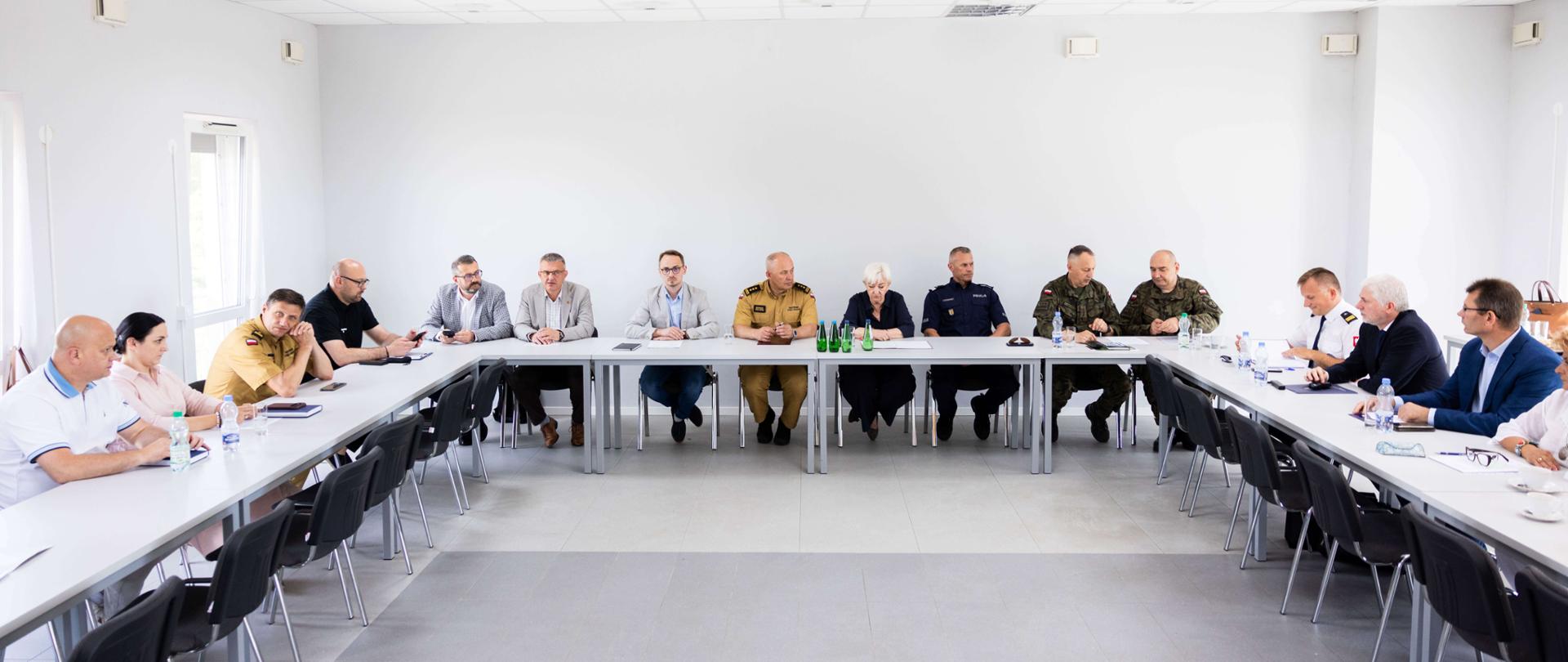 Oficjalne spotkanie grupy osób siedzących przy stołach ustawionych w podkowę w jasnej sali konferencyjnej. Wśród uczestników widoczni są przedstawiciele różnych służb mundurowych w oficjalnych mundurach (straż pożarna, wojsko, policja) oraz osoby w strojach cywilnych.